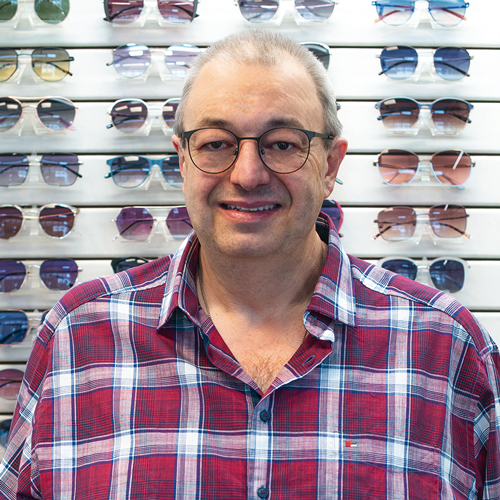 Mann mit rotem Hemd und einer Brille steht vor einer Wand mit Sonnenbrillen.