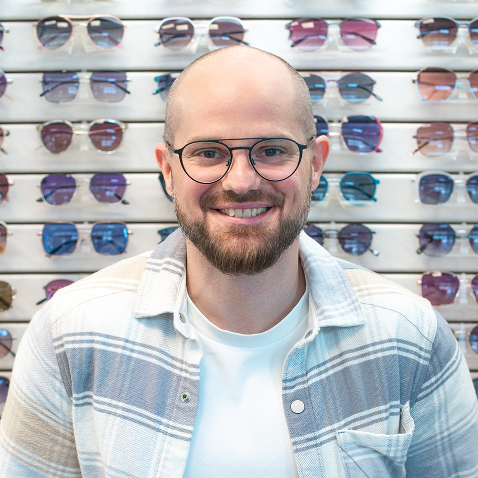 Mann mit Hemd und einer Brille steht vor einer Wand mit Sonnenbrillen.
